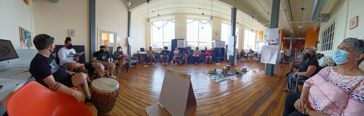 A group of people sitting in a circle in a sunny room. In the middle of the circle are various objects - drums, flip chart paper, a cloth with items on it. There are a few columns in the room with flip charts stuck to them.